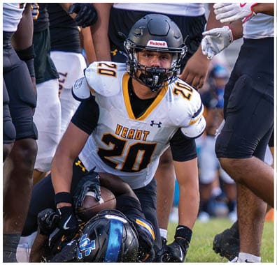 A person wearing a white football jersey with the number 20 and a black helmet is holding a football while standing over another person lying on the ground during a game. Descripci n generada con IA