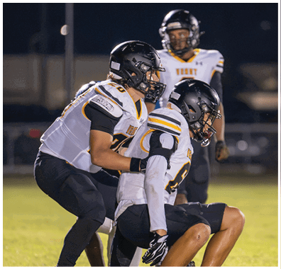 Two people in football uniforms and helmets are engaged in a play on a field at night. Descripci n generada con IA
