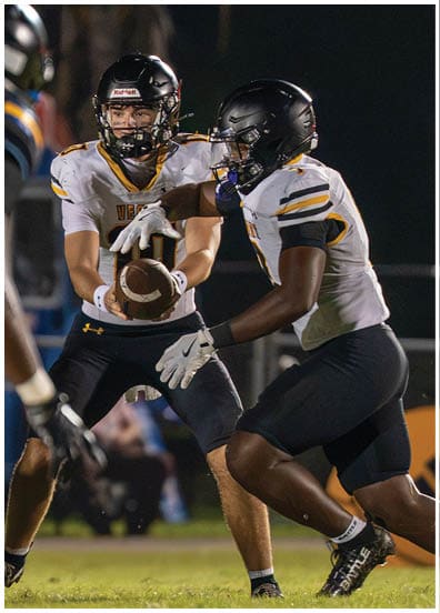 Two people in football uniforms are engaged in a play with one person handing off the football to the other on a nighttime field. Descripci n generada con IA