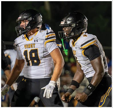 Four people wearing football uniforms and helmets are on a grassy field at night, with one person holding a football and the others appearing to prepare for a play. Descripci n generada con IA