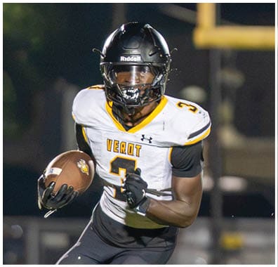 A person wearing a black helmet and white sports jersey with the number 3 is running with a football while another person in a dark helmet and sports uniform approaches from the right on a grassy field at night. Descripci n generada con IA
