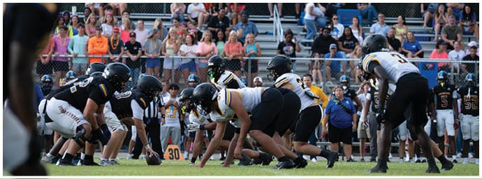 Two opposing American football teams in helmets and uniforms are lined up facing each other on a field with a crowd of people watching from the bleachers behind a chain-link fence. Descripci n generada con IA