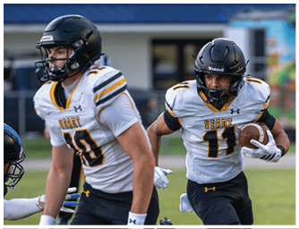 Two people wearing football helmets and uniforms are actively playing on a grassy field, with one holding a football and running while the other is nearby. Descripci n generada con IA
