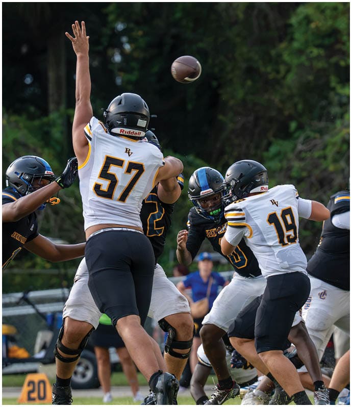 People wearing football helmets and uniforms are engaged in a play on a grassy field with a blurred person in the background. Descripci n generada con IA