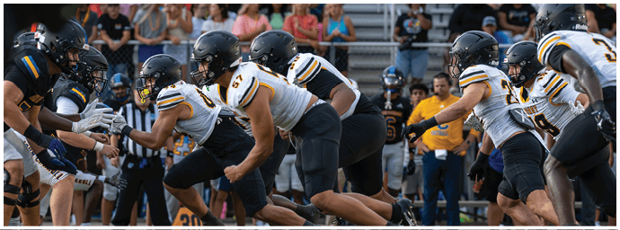 Two opposing American football teams are lined up facing each other on the field, preparing for the next play with players in helmets and uniforms. Descripci n generada con IA