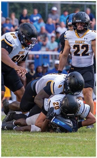Two people in football uniforms and helmets are engaged in a play on a field with a blurred crowd in the background. Descripci n generada con IA