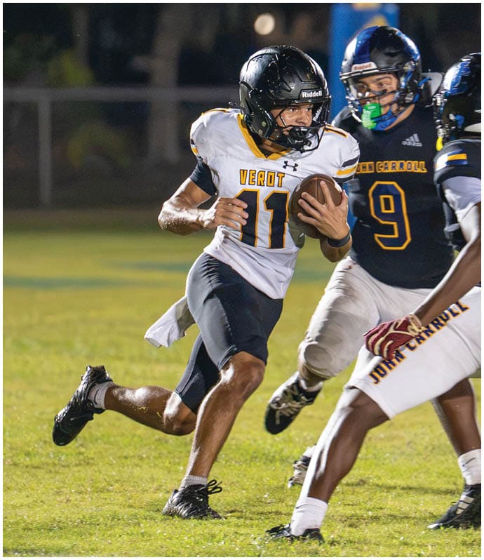 A person wearing a white and black football uniform is running with the ball while two people in dark blue and white football uniforms attempt to block or tackle them on a grassy field at night. Descripci n generada con IA