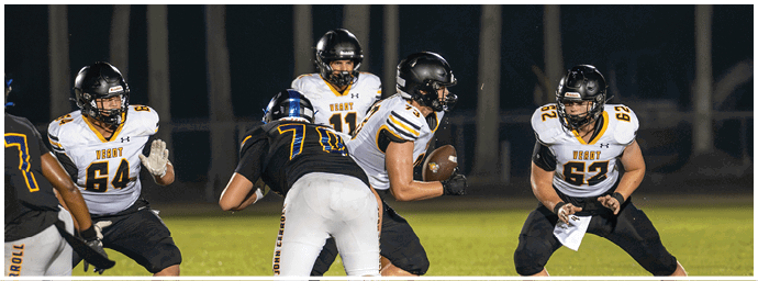 People wearing football uniforms are engaged in a play on a grassy field at night with one person holding the ball and others preparing to block or tackle. Descripci n generada con IA