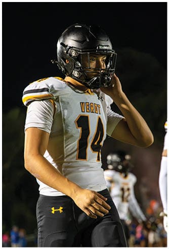 A person wearing a football helmet and a white jersey with the number 14 and the word “VEROT" stands on a field at night. Descripci n generada con IA