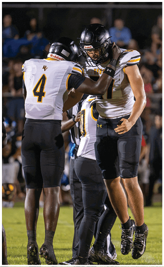 Two people wearing football helmets and white jerseys with black and yellow accents are engaged in a close interaction on a field. Descripci n generada con IA