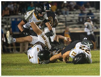 Several people wearing football uniforms are engaged in a tackle on a grassy field during a game with blurred spectators in the background. Descripci n generada con IA