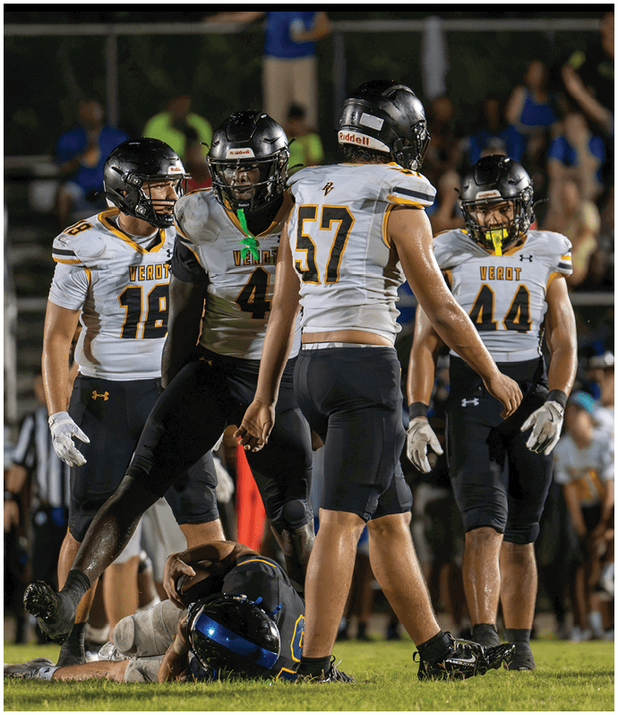 Four football players in white and black uniforms with helmets stand on a field while one player in a blue and yellow uniform lies on the ground. Descripci n generada con IA