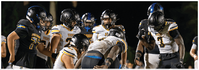 People wearing football uniforms and helmets are gathered closely on a field under bright lights during a nighttime game. Descripci n generada con IA