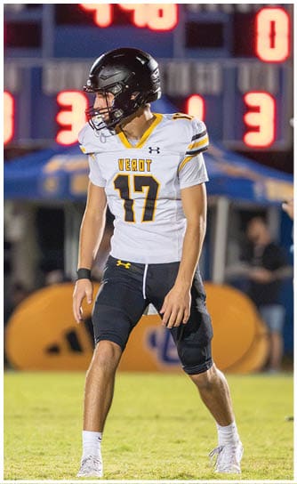 A person wearing a football uniform with the number 17 stands on a grassy field during a game with a scoreboard visible in the background. Descripci n generada con IA