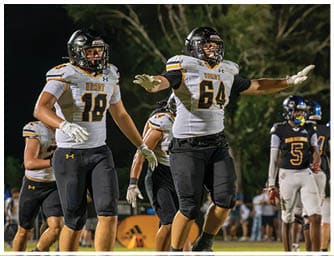 Two people in football uniforms with helmets are on a field at night, one with arms extended and the other standing nearby, while other players and spectators are visible in the background. Descripci n generada con IA