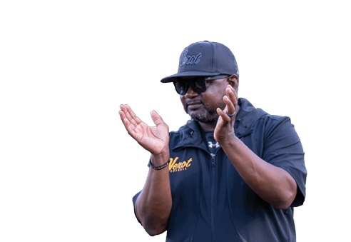 A person wearing a black cap, sunglasses, a black sleeveless jacket, light-colored pants, and black shoes is standing on grass and clapping. Descripci n generada con IA