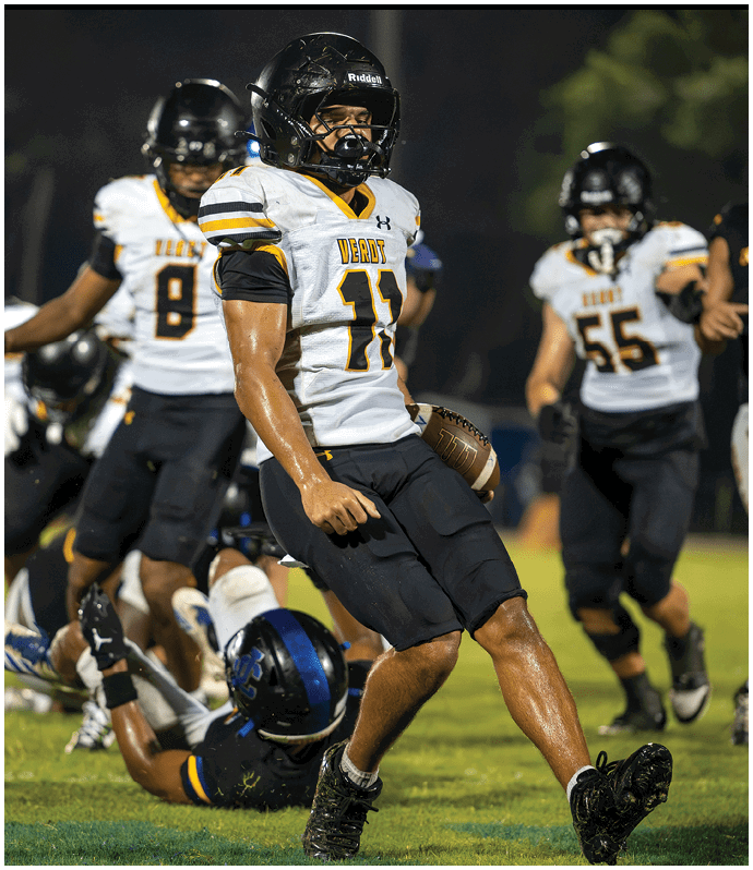 A person wearing a football uniform with the number 11 is holding a football while another player is on the ground and others are running in the background on a grassy field. Descripci n generada con IA