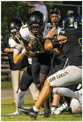 Two football players in black helmets and white jerseys are engaged in a play on a grassy field with a blurred background of people and bleachers. Descripci n generada con IA