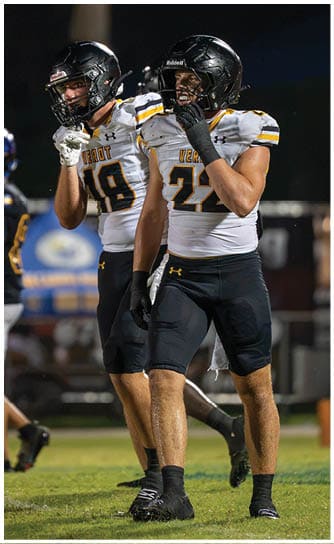 Two adults wearing football helmets and white jerseys with black and yellow accents stand on a field at night, with one person touching their faceguard. Descripci n generada con IA