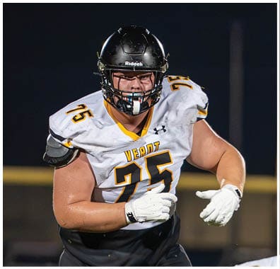 A person wearing a black football helmet and a white jersey with yellow and black details is focused and appears to be in action during a game. Descripci n generada con IA