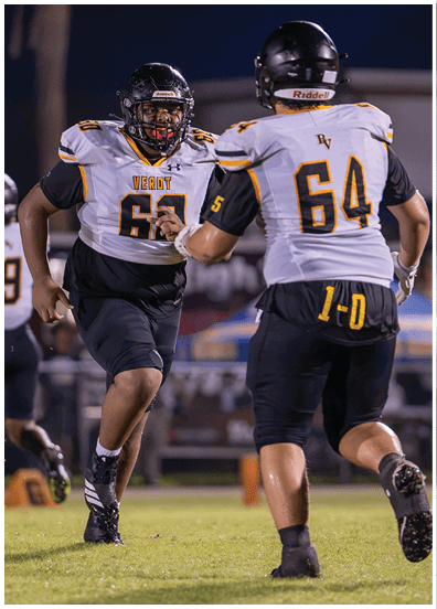 Two people wearing football helmets and white jerseys with black and yellow accents are engaged in a play on a football field at night. Descripci n generada con IA