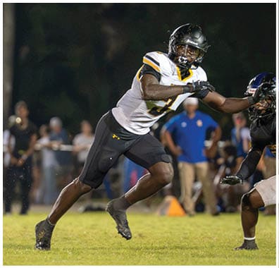 Two football players in action on a field with one player in a white and black uniform reaching out to block or tackle the other player in a black and yellow uniform holding the ball. Descripci n generada con IA