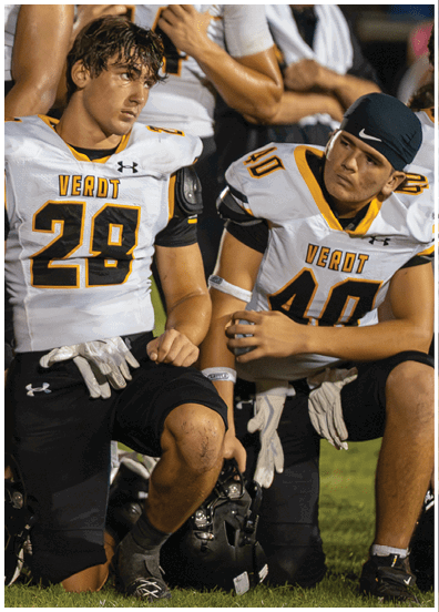 Four people wearing football uniforms with the word “VERDT" on the jerseys are kneeling on grass, holding helmets and appearing focused or attentive. Descripci n generada con IA