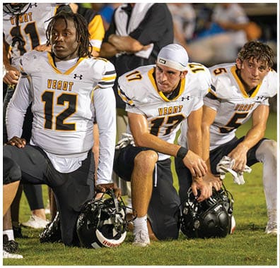Two athletes in white football uniforms with “VEROT" on the front are on the sidelines, one standing and the other sitting with a focused expression. Descripci n generada con IA