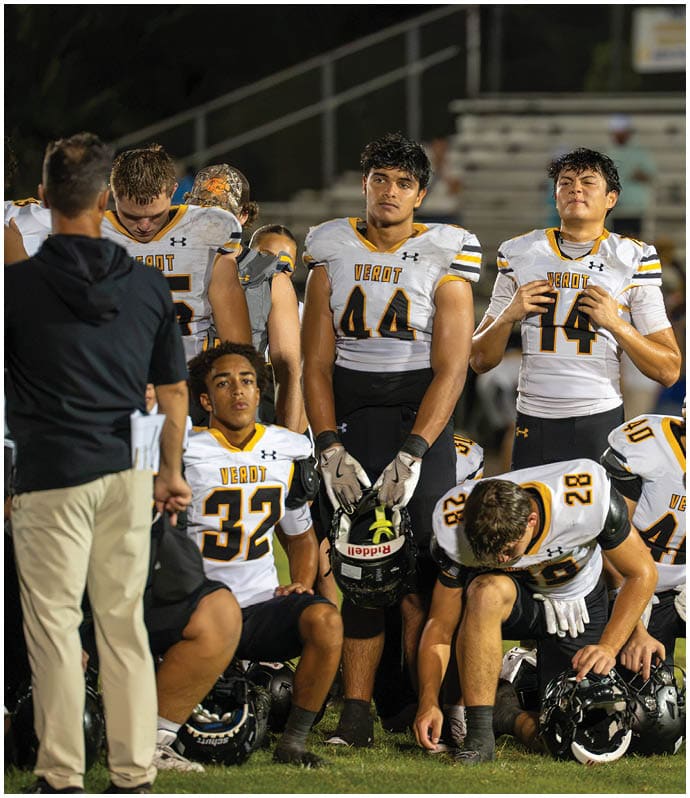 A group of football players in white and yellow uniforms are gathered on a field, some standing and some sitting, with a coach or staff member nearby and empty bleachers in the background. Descripci n generada con IA