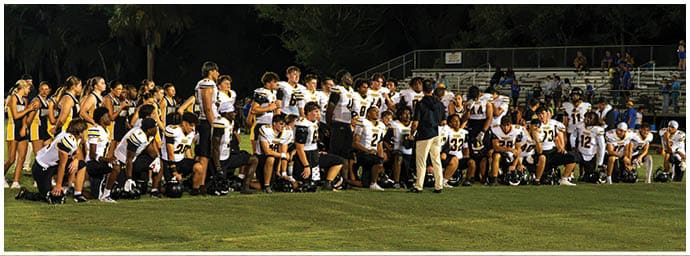 A large group of people wearing sports uniforms and coaches are gathered on a grassy field at night with some people sitting in bleachers in the background. Descripci n generada con IA