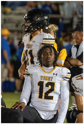 A group of people wearing football uniforms with “VEROT" on the jerseys are gathered on a field, some standing and some sitting, appearing focused and contemplative during a game or practice. Descripci n generada con IA