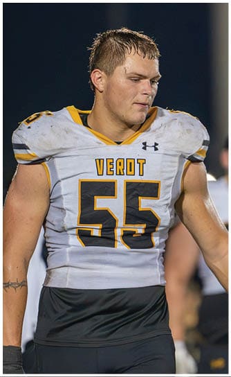 An adult wearing a white and black football uniform with the number 55 and the word Verot on the chest stands on a field at night with a focused expression. Descripci n generada con IA