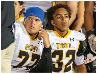 Four adults in football uniforms sitting on the grass with helmets on the ground, appearing attentive or contemplative during a game or practice. Descripci n generada con IA