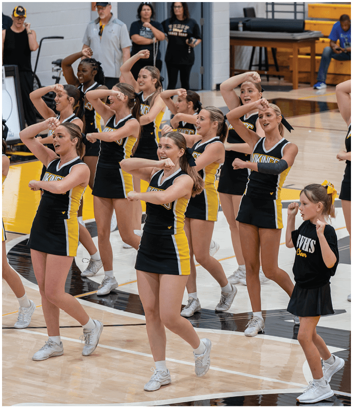 A group of adults and a child in cheerleading uniforms are performing a synchronized routine on a gymnasium floor with spectators in the background. Descripci n generada con IA