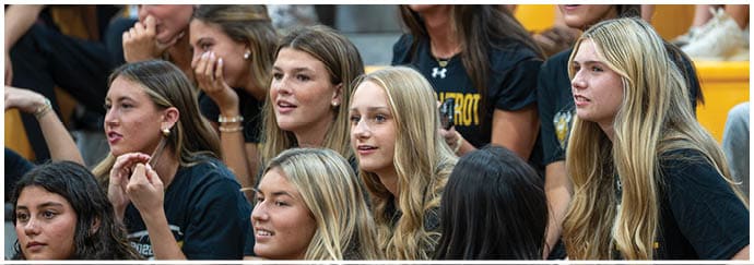 A group of people with long hair are sitting closely together, some smiling and others looking attentively in the same direction, wearing casual clothing in a bright indoor setting. Descripci n generada con IA