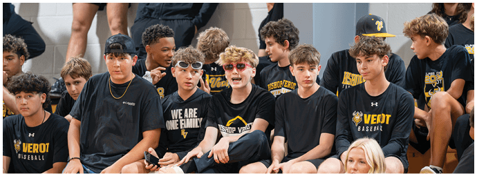 A group of people wearing black and yellow sports-themed clothing are sitting closely together against a wall, some smiling and engaging with each other. Descripci n generada con IA