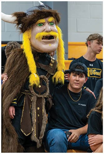 A person is wearing a large Viking-style costume head with bright yellow braided hair and mustache, along with a fur cloak and dark clothing with gold accents. Descripci n generada con IA