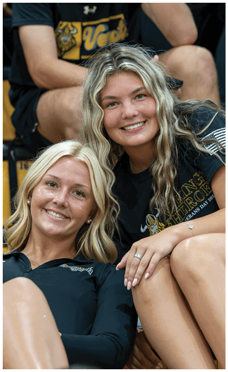 Two smiling adults with wavy hair are sitting close together wearing black shirts, one with a graphic design and the other with text. Descripci n generada con IA