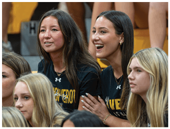 Two adults with long hair wearing black shirts are sitting closely together, one smiling and the other looking ahead, in a setting with yellow seats. Descripci n generada con IA