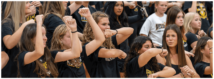 A group of people wearing matching black shirts are enthusiastically raising their arms and cheering in what appears to be a sports event or pep rally. Descripci n generada con IA
