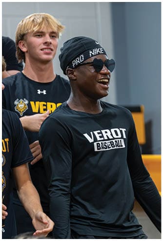 A person wearing heart-shaped sunglasses, a black Nike headband, and a black long-sleeve shirt with “VEROT BASEBALL" printed on it is smiling and standing indoors near yellow seating. Descripci n generada con IA