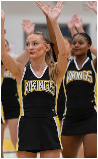 A group of adults in black and yellow cheerleading uniforms with the word Vikings are raising their arms in a gymnasium. Descripci n generada con IA