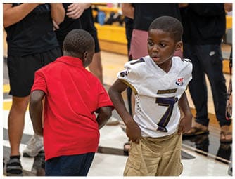 Two children stand indoors on a gym floor, one wearing a white sports jersey and khaki shorts and the other in a red shirt and dark shorts. Descripci n generada con IA