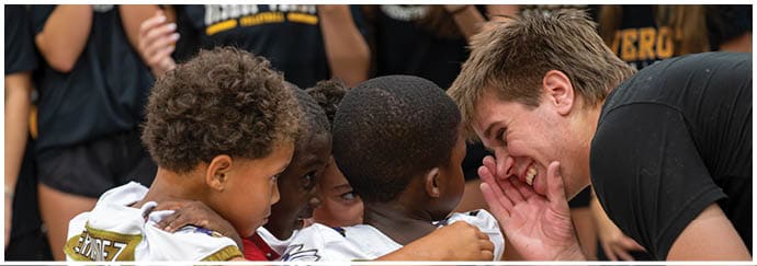 An adult is crouched down speaking closely and smiling to a group of children wearing sports jerseys who have their arms around each other. Descripci n generada con IA