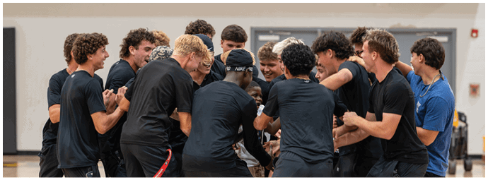 A group of people wearing athletic clothing are gathered closely in a gymnasium, appearing to celebrate or cheer together. Descripci n generada con IA