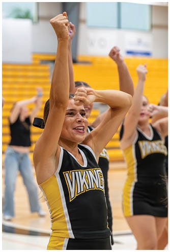 A person wearing a Vikings cheerleading uniform is raising one arm enthusiastically in a gymnasium with yellow bleachers in the background. Descripci n generada con IA