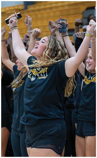 A group of people wearing matching black sports shirts and shorts are raising their arms and cheering enthusiastically in an indoor gymnasium. Descripci n generada con IA