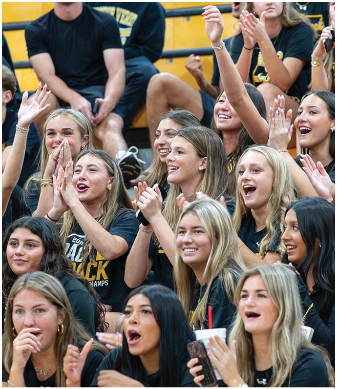 A group of people wearing matching black shirts are enthusiastically cheering and clapping in what appears to be a sports or school event. Descripci n generada con IA