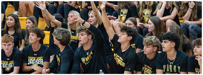 A group of people wearing matching black shirts with yellow text are sitting closely together on bleachers, with some raising their hands and others looking in various directions. Descripci n generada con IA