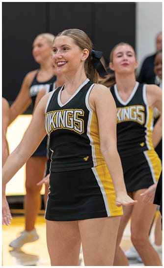 Adults wearing black and yellow cheerleading uniforms with the word Vikings perform a routine indoors. Descripci n generada con IA